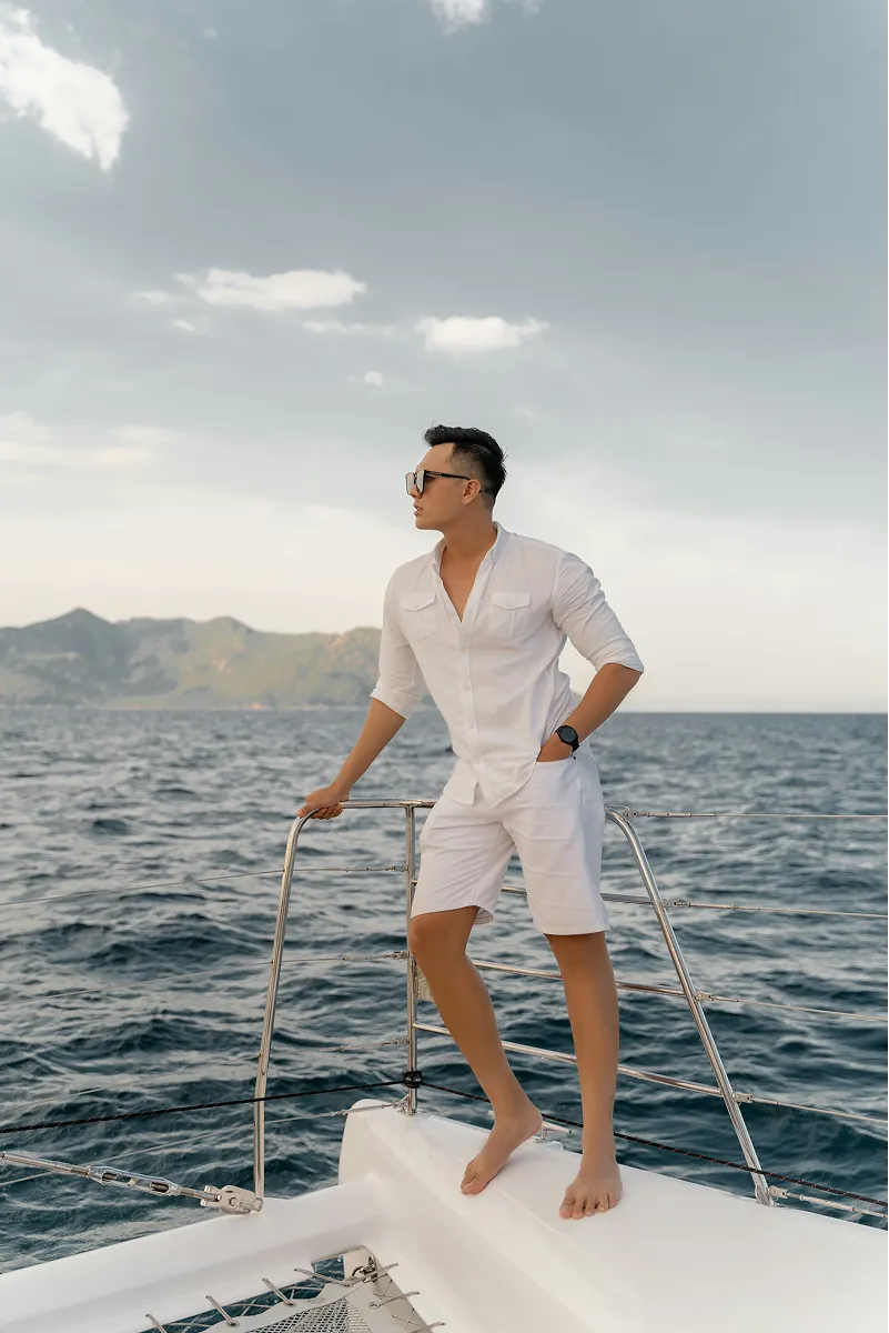 A man standing on the bow rail of a yacht, looking out to a headland.